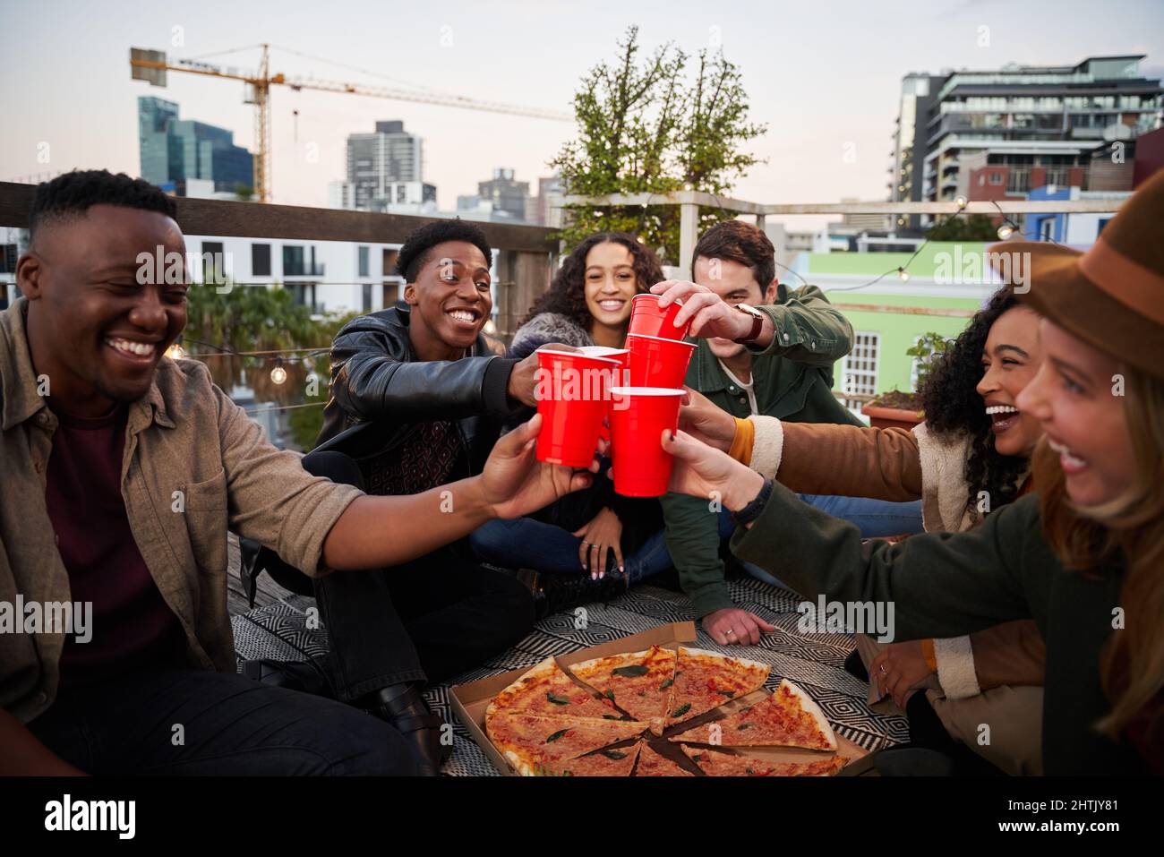 Gruppo di amici diversi che fanno un brindisi con tazze rosse mentre si siede sulla terrazza sul tetto della città. Bei tempi e felicità Foto Stock