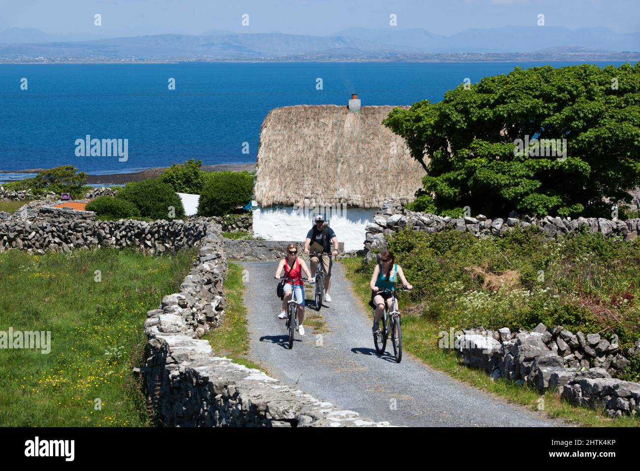 Cottage dipinto di bianco con tetto di paglia con ciclisti tra pareti di pietra asciutta e Galway Bay dietro, Inishmore, Aran Islands, County Galway, Irlanda Foto Stock