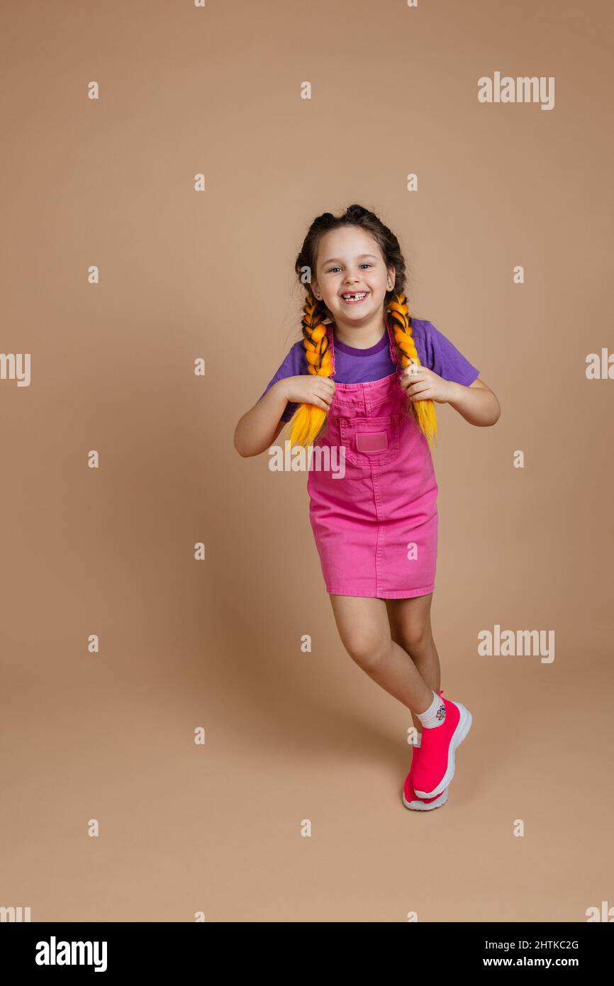 Commovente, gioiosa, bambina che tocca i pigtail kanekalon giocando, ballando in un posto guardando la macchina fotografica, indulgere sorridendo con il dente mancante in rosa Foto Stock