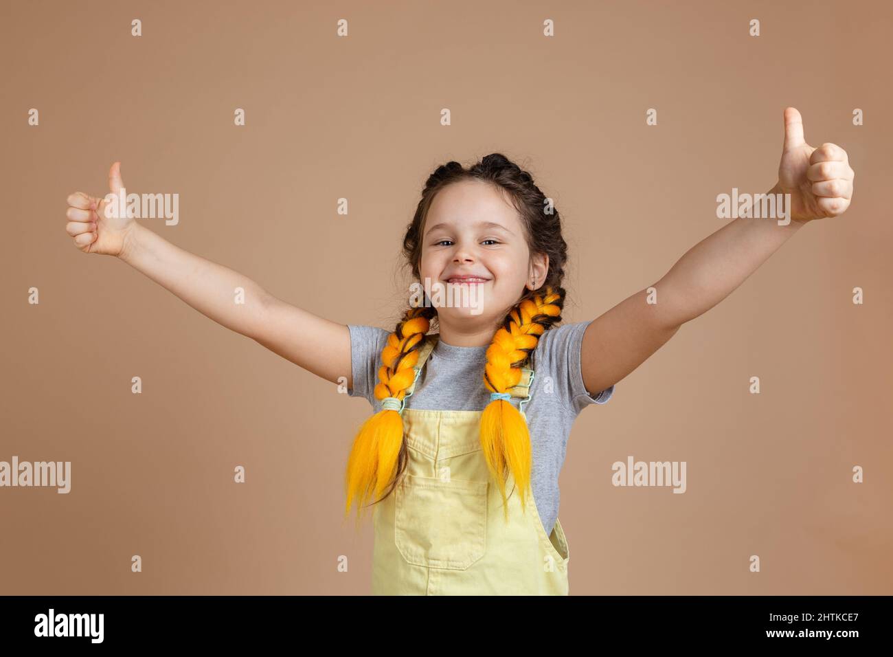 Giovane ragazza sorridente felice con pigtail giallo kanekalon guardando la macchina fotografica, mostrando come i segni con le mani indossare tuta gialla e t grigio Foto Stock