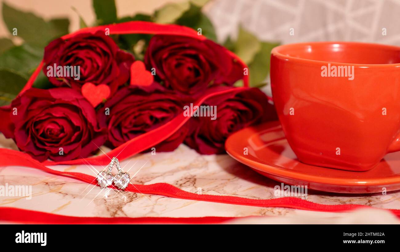 Sfondo festivo Flatlay, composizione per San Valentino il 14 febbraio. Caffè in una tazza rossa, fiori di rosa rossa e orecchini a cuore di diamante a casa a letto Foto Stock