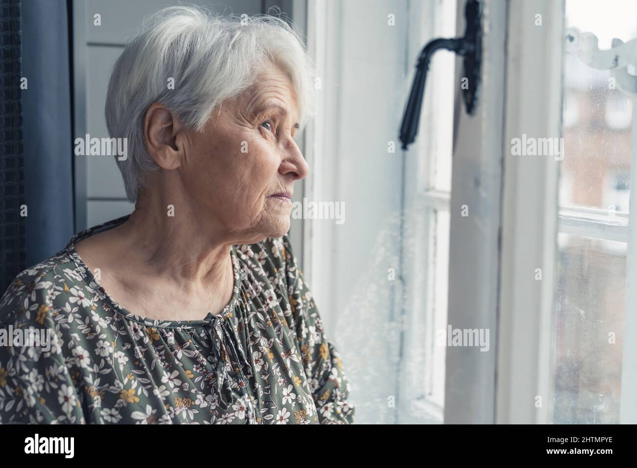 signora di pensionato caucasico dai capelli grigi che guarda tristemente dalla finestra medio primo piano in su colpo interno. Foto di alta qualità Foto Stock