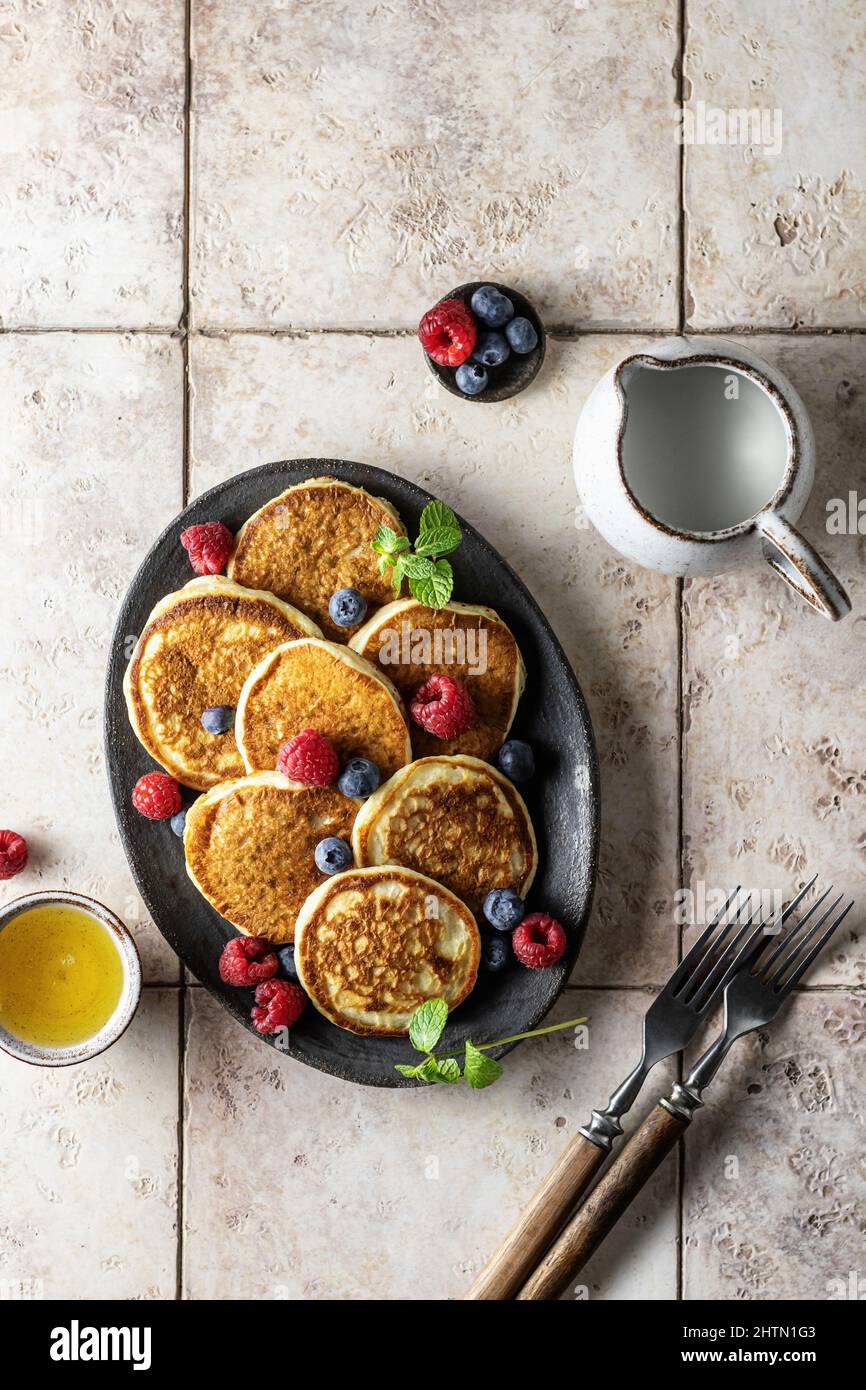 Pancake in piastra di ceramica con lamponi, mirtilli, foglie di menta, forchette su sfondo di tegole rosa, vista dall'alto Foto Stock