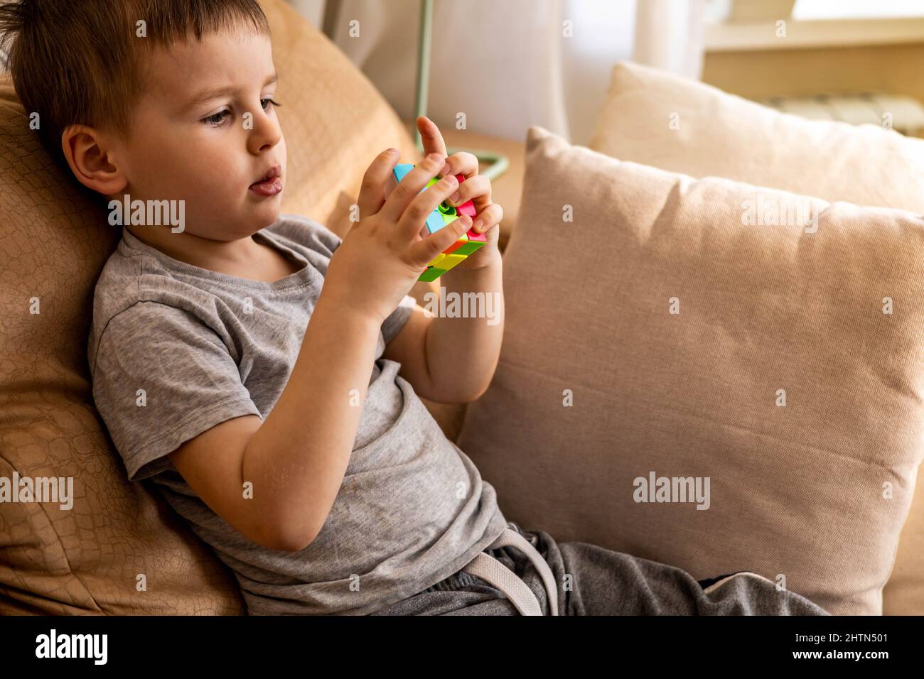 Bambino ragazzo carino che monta cubo multicolore di Rubik risolvere gioco logico che si trova sul divano a casa Foto Stock
