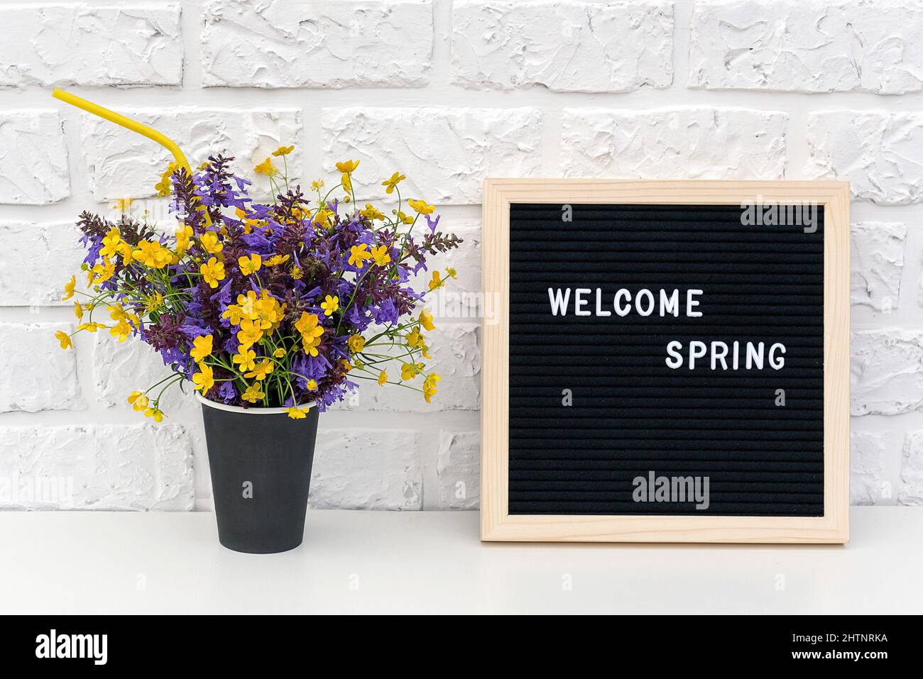 Testo Ciao primavera su lavagna nera e bouquet di fiori colorati in carta nera tazza di caffè con cannuccia cocktail su sfondo bianco mattone parete. C Foto Stock