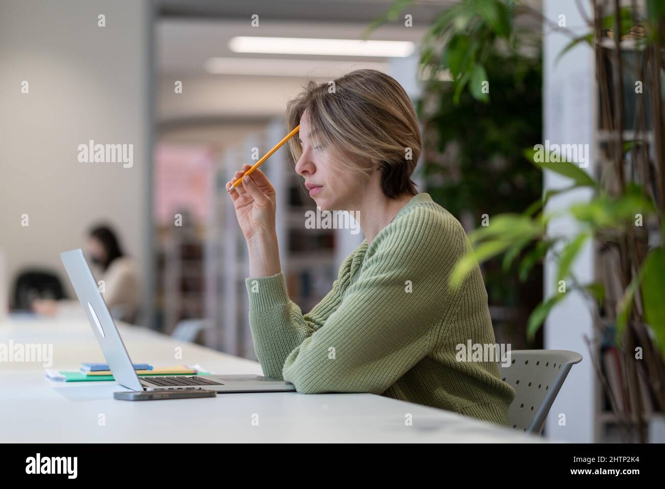 Studentessa matura femminile concentrata che studia online su laptop in una moderna biblioteca circondata dal verde Foto Stock