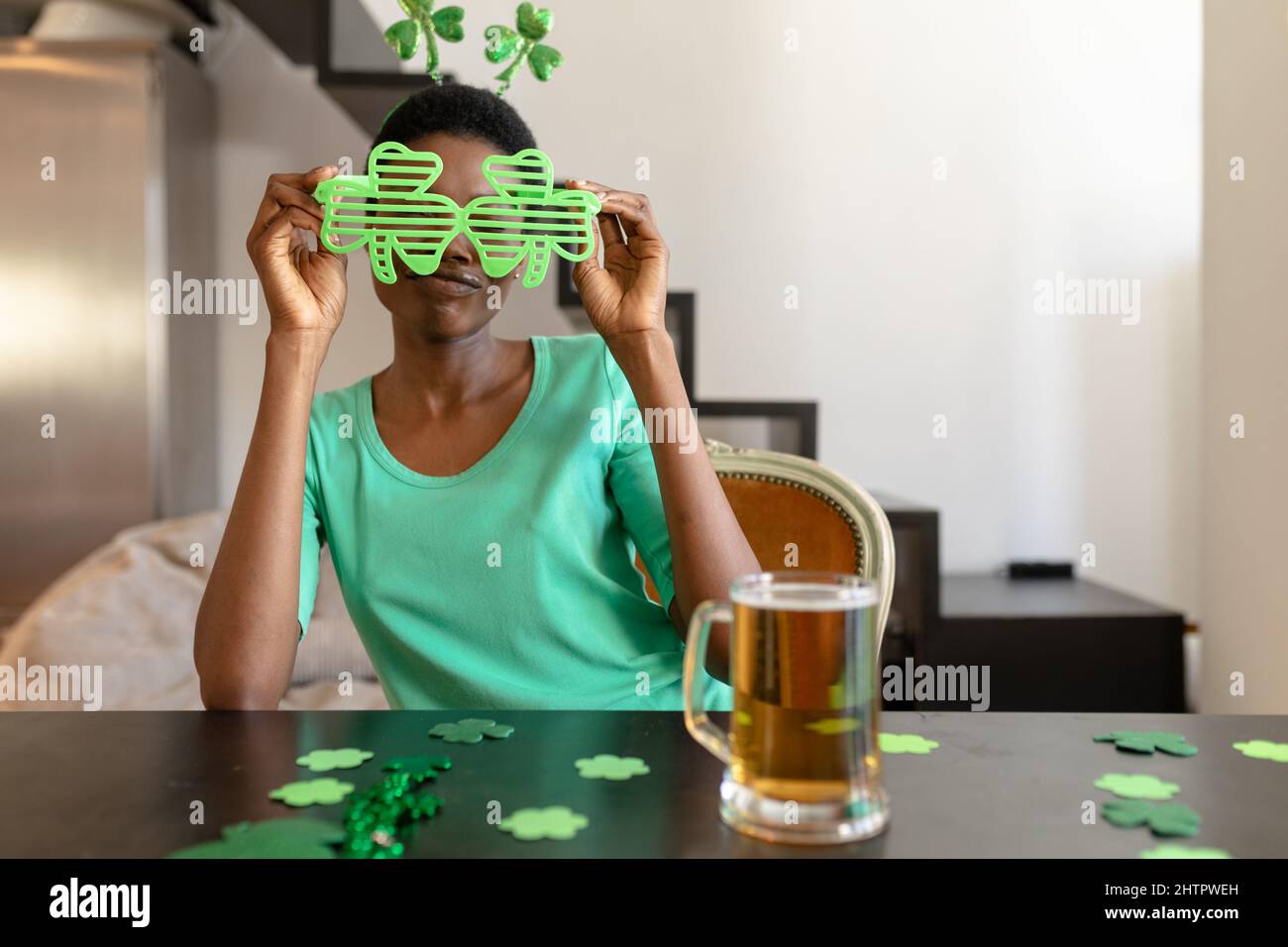 Donna africana americana che tiene shamrock occhiali di novità mentre si siede con tazza di birra al tavolo Foto Stock