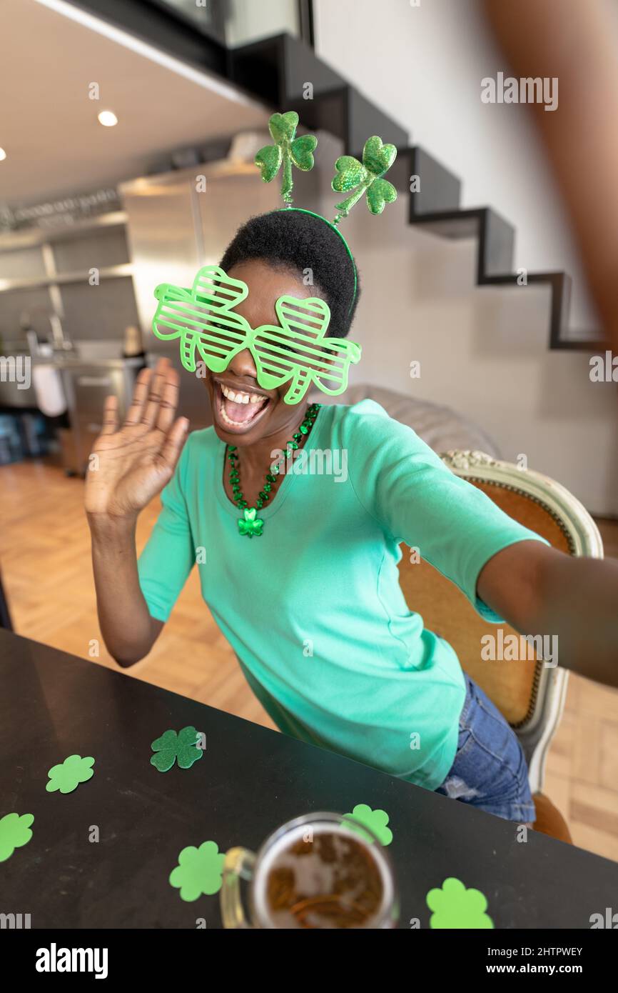 Donna afro-americana felice che indossa occhiali shamrock novità mentre prende selfie a casa Foto Stock