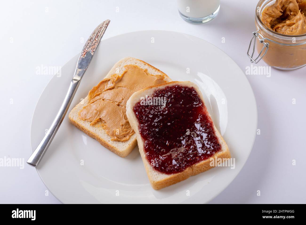 Primo piano di burro di arachidi e conserva su fette di pane nel piatto con coltello da tavolo, vasetto e latte Foto Stock