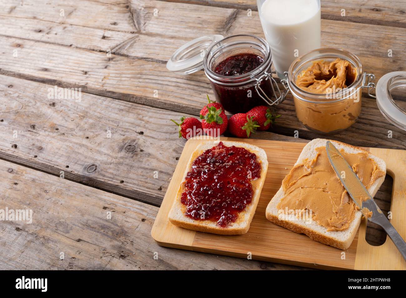 Panino con burro di arachidi e gelatina a bordo con vasetti, fragole e latte a tavola Foto Stock