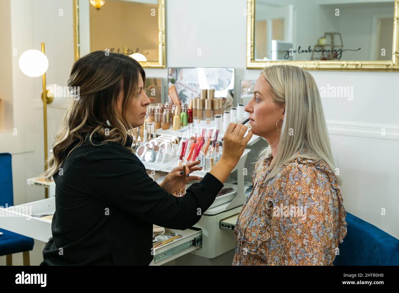 Cosmetologist che applica il trucco del rivestimento del labbro ad una donna bionda in una sedia di trucco Foto Stock