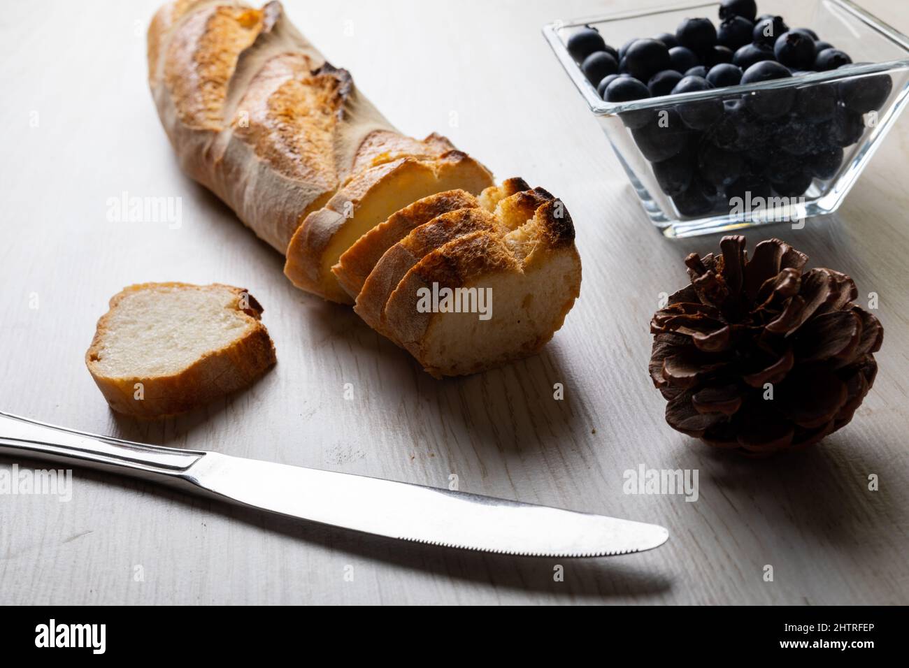 Primo piano di baguette affettate con cono di pino e coltello da tavolo con mirtilli in ciotola sul tavolo Foto Stock