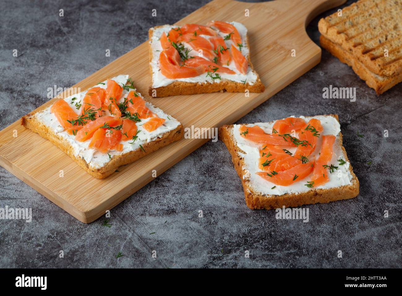 Pane fatto in casa su un tagliere di legno con formaggio di cagliata, salmone ed erbe Foto Stock