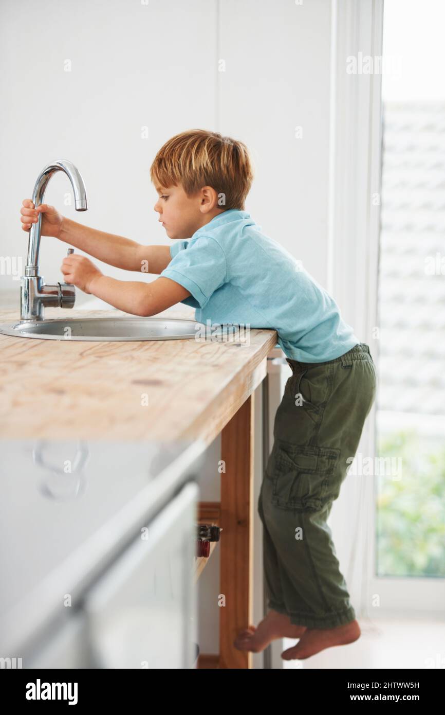 Voglio avere un po' d'acqua.... Un ragazzo giovane carino che sale sul banco della cucina per arrivare al rubinetto. Foto Stock