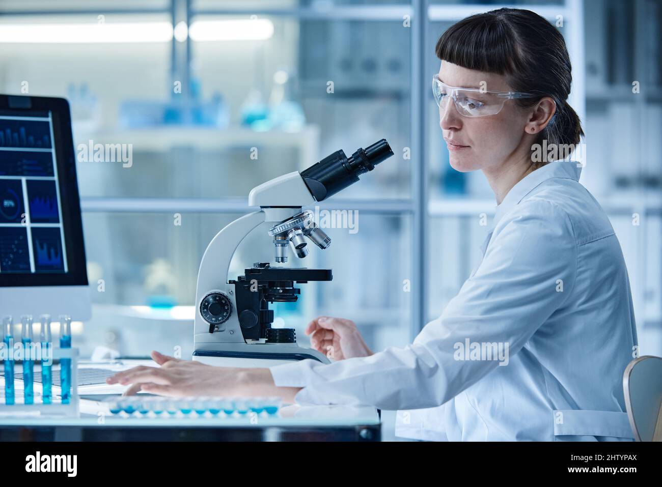 Scienziata femminile seduta al tavolo in laboratorio ed esame dei campioni in provette con microscopio Foto Stock