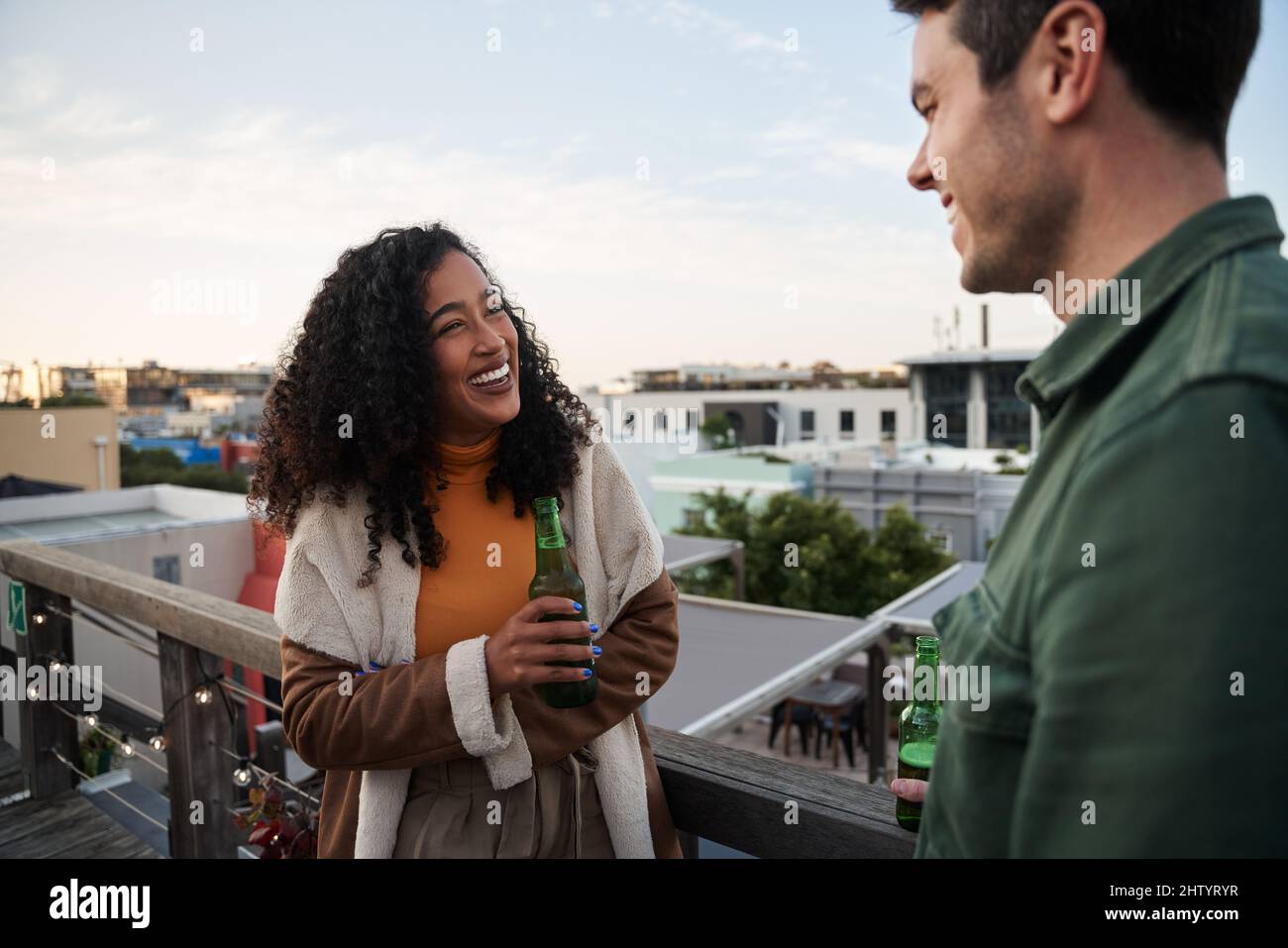 Biracial giovane adulta femmina ridendo con un amico sul balcone di una terrazza sul tetto della città. Drink a portata di mano, vista sulla città Foto Stock