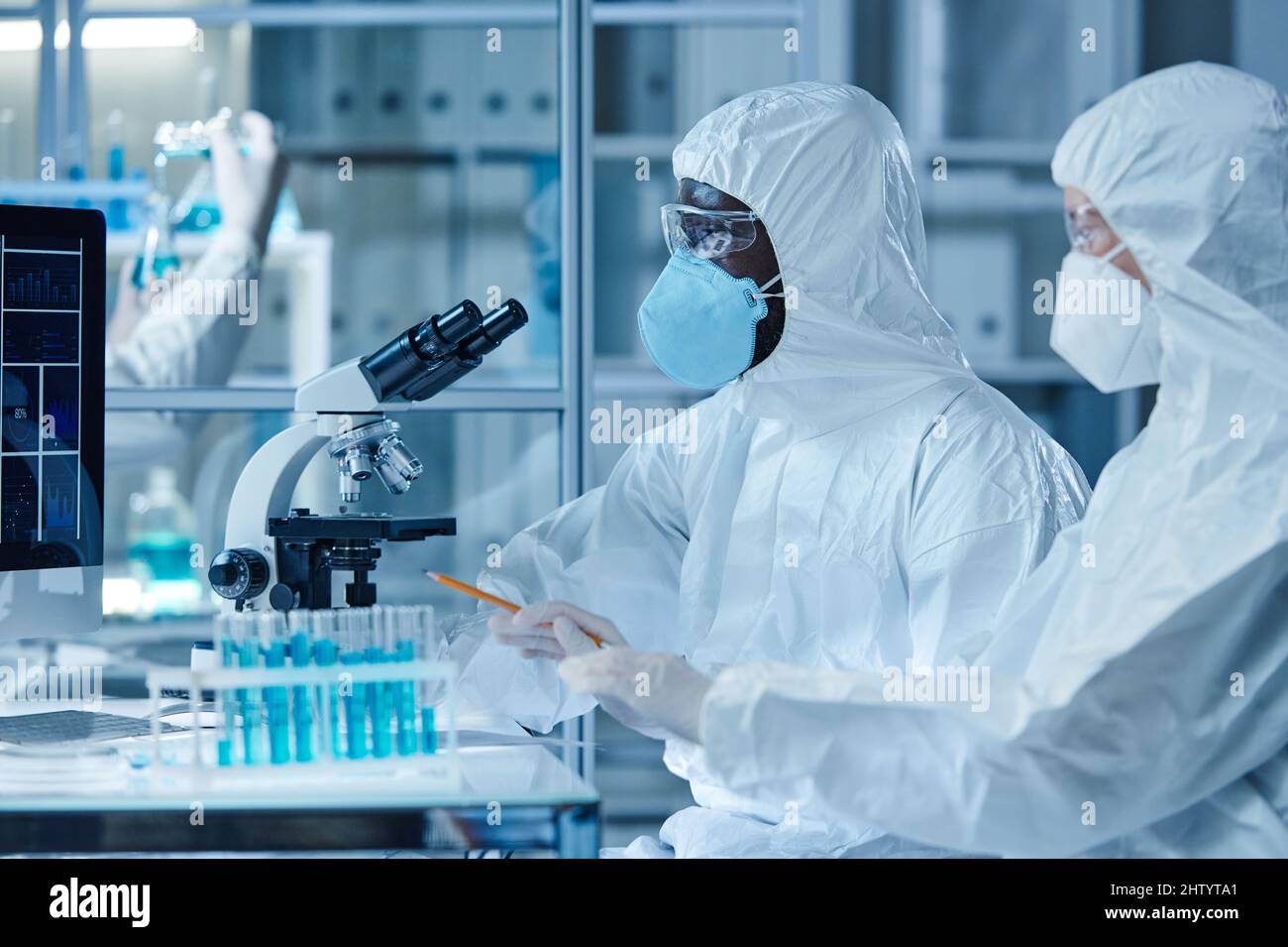 Due scienziati in tute protettive che lavorano con l'analisi in provette mentre sono seduti al tavolo con microscopio in laboratorio Foto Stock