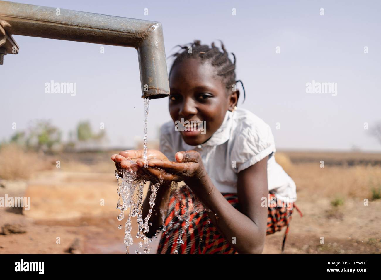 Ragazza africana nera intelligente seduta di fronte ad un rubinetto che beve acqua fresca; concetto di scarsità d'acqua Foto Stock