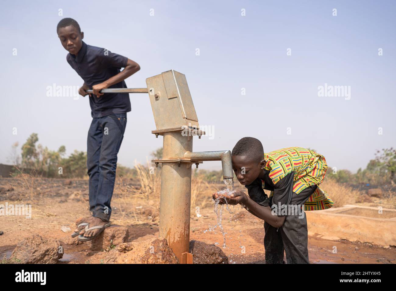 Un diligente giovane nero che pompa acqua potabile fresca per il suo fratellino assetato in un rubinetto manuale pubblico in qualche parte dell'Africa occidentale Foto Stock