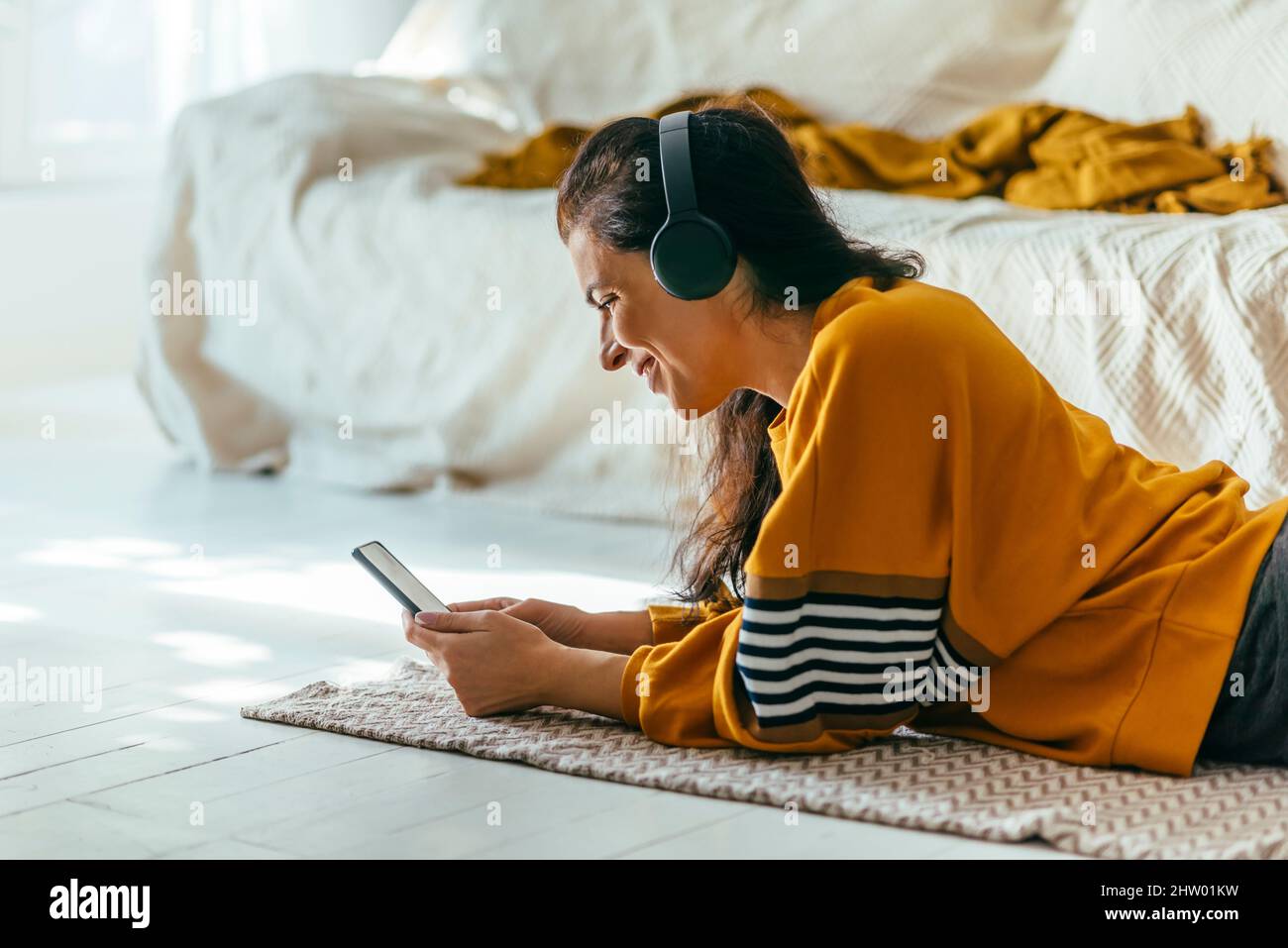 Donna felice in casual abbigliamento giallo sdraiato giù pavimento e utilizzando il telefono per videochiamata a casa sua. Persona che ha tempo libero al coperto guardando smartp Foto Stock