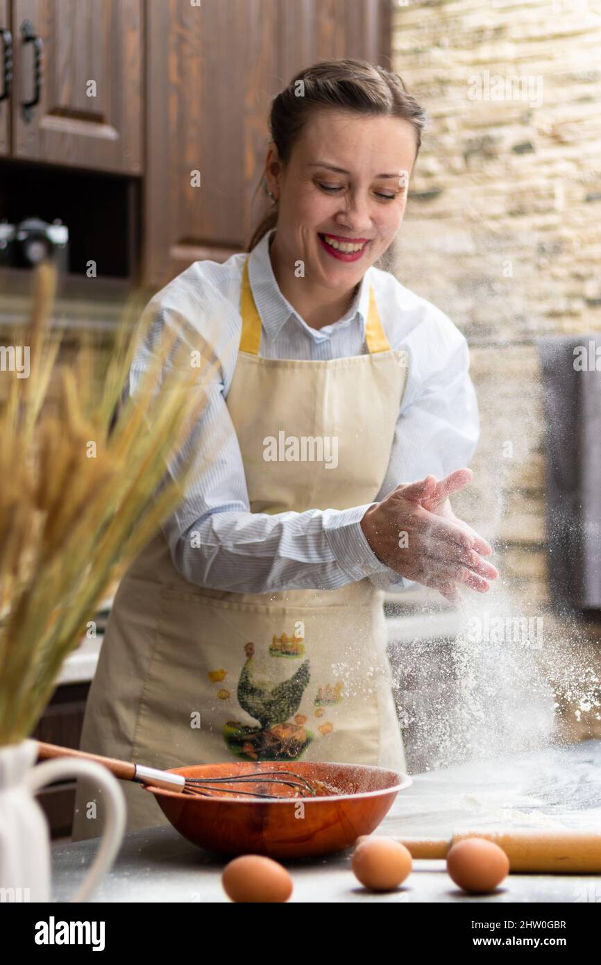 Una giovane e bella cuoca in un grembiule indulge in farina a casa in cucina sullo sfondo degli utensili da cucina. Su un tavolo di legno sono ad es Foto Stock