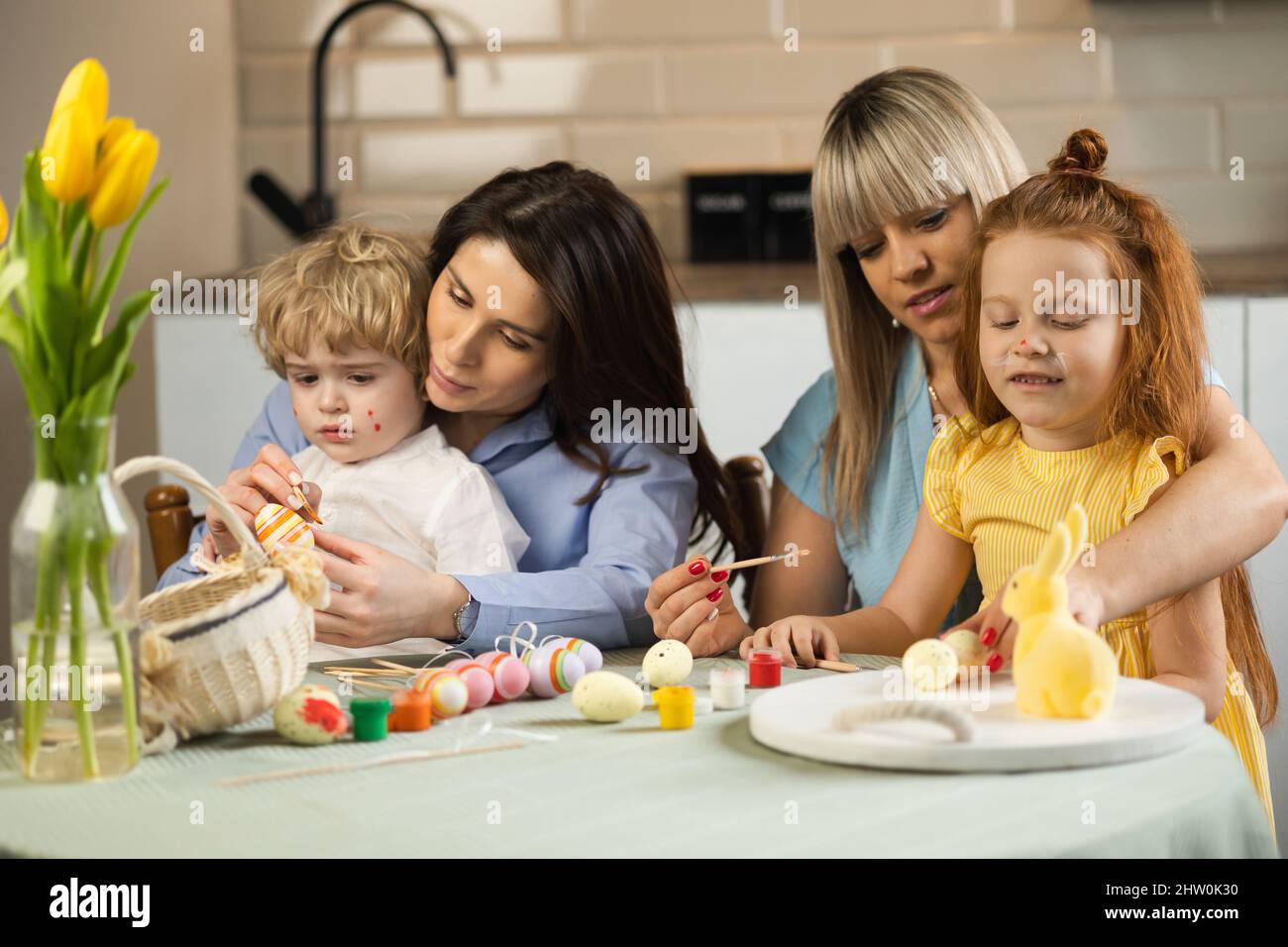 Due madri con i loro figli dipingono uova di Pasqua in cucina Foto Stock