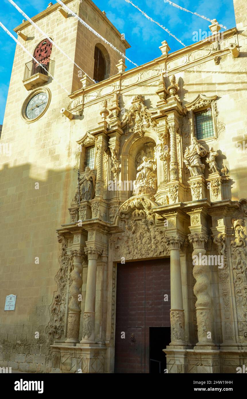 Ingresso artistico della Basilica di Santa Maria di Alicante tra la torre dell'orologio e un'altra torre sotto il cielo azzurro. Stile gotico valenciano dal Foto Stock