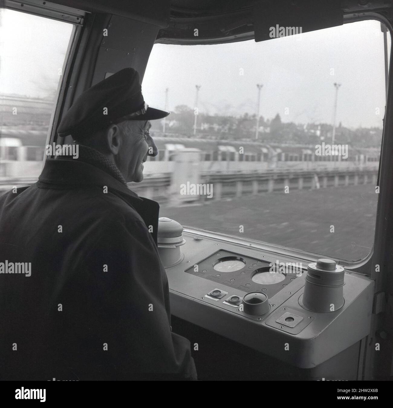 1960s, storico, un driver della metropolitana in berretto e giacca, seduto nella cabina del conducente di un treno della metropolitana di Londra, Londra, Inghilterra, Regno Unito. Noti come stock del 1967, i treni operarono per la prima volta sulla linea Victoria, aperta di recente, nel 1968 e furono in uso fino al 2011. Dotati del sistema Automatic Train Operation (ATO), sono stati i primi treni ferroviari automatici al mondo. Sono stati anche il primo materiale rotabile della metropolitana di Londra a presentare finestre avvolgenti nella cabina del conducente. Foto Stock