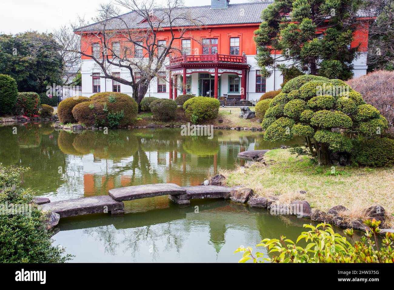 Fantastico boxwood topiary albero a piccola isola e ponte e stagno pietra e un po 'di edificio rosso. All'angolo sinistro il cespuglio camelia con fiori rossi - Koshika Foto Stock