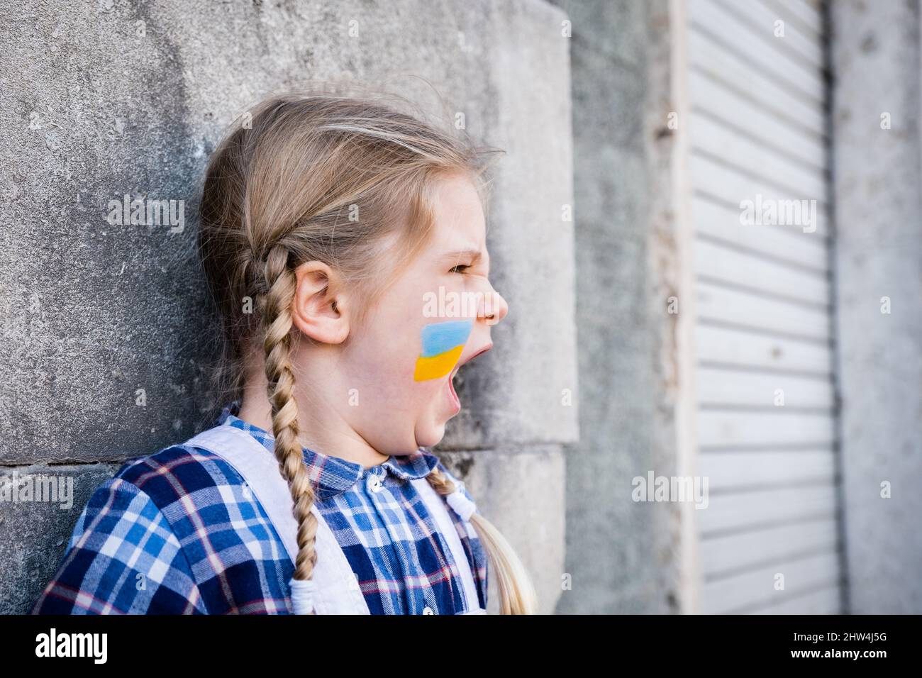 Ritratto di una bambina che grida e con bandiera dipinta sulla guancia nei colori giallo-blu della bandiera Ucraina. Concetto di pace e pr Foto Stock