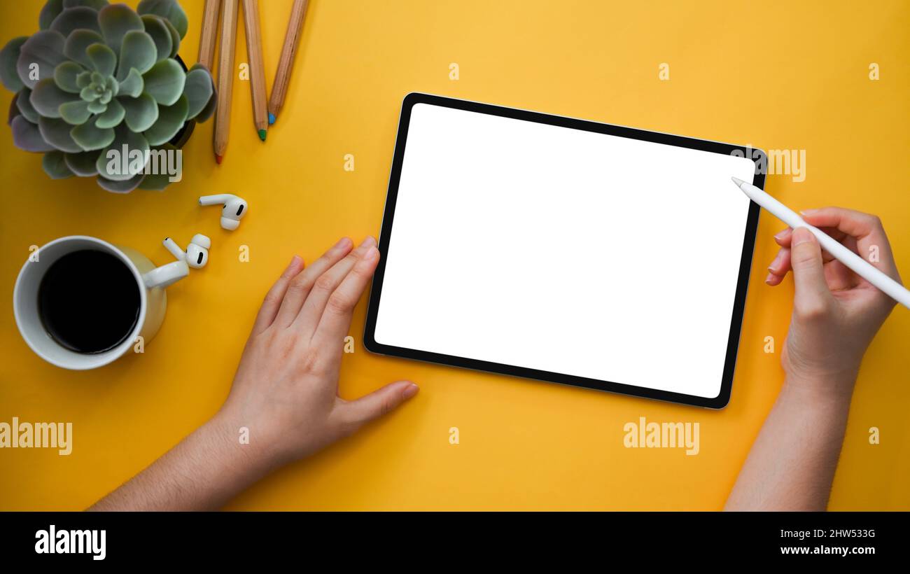 Elegante sfondo giallo della scrivania da ufficio con una mano femminile che disegna grafica su un mockup schermo bianco del touchpad del tablet digitale. Vista dall'alto Foto Stock