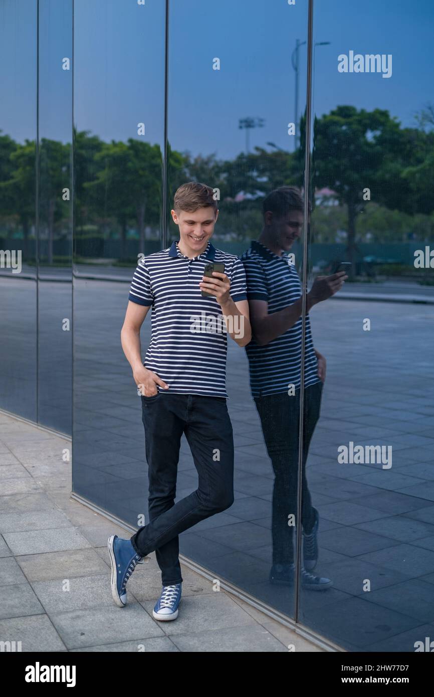 Giovane uomo caucasico in abbigliamento casual in piedi vicino moderno edificio di vetro e guardando il suo telefono. Persone che utilizzano telefoni. Foto di alta qualità Foto Stock