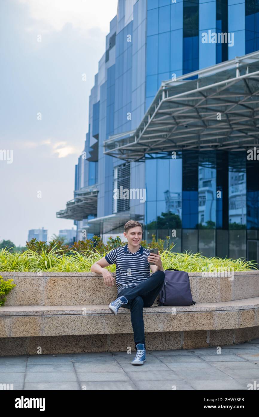 Giovane uomo caucasico in abbigliamento casual seduto vicino al moderno edificio di uffici e fare videochiamata. Foto di alta qualità Foto Stock