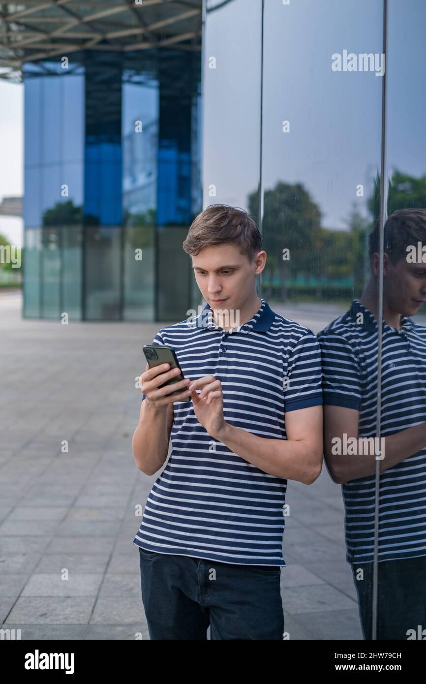 Giovane uomo caucasico in abbigliamento casual in piedi vicino moderno edificio di vetro e guardando il suo telefono. Persone che utilizzano telefoni. Foto di alta qualità Foto Stock