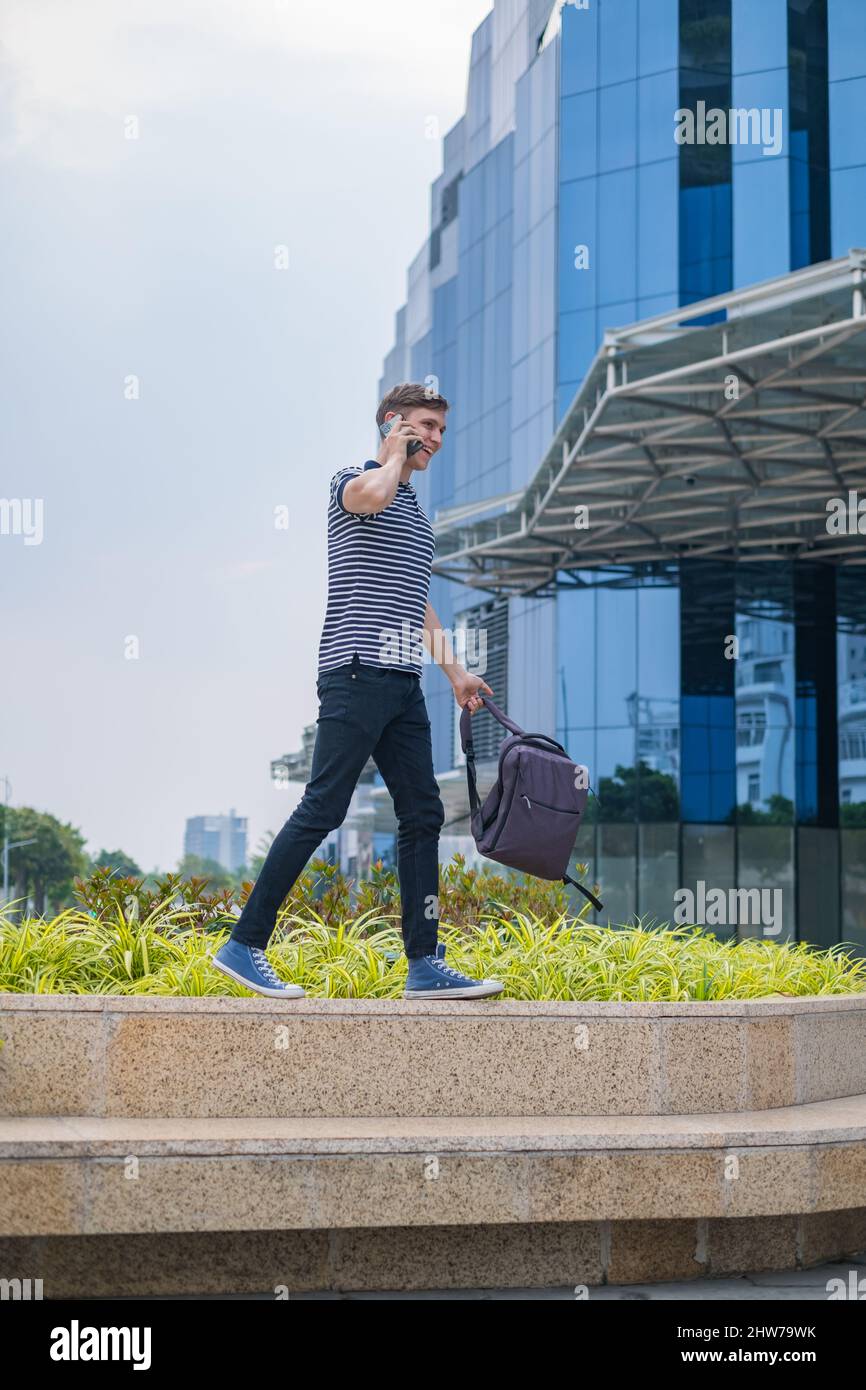 Giovane uomo caucasico in abbigliamento casual con lo zaino che cammina vicino a moderni edifici d'ufficio e fare telefonate. Persone che usano il telefono. Alta qualità Foto Stock