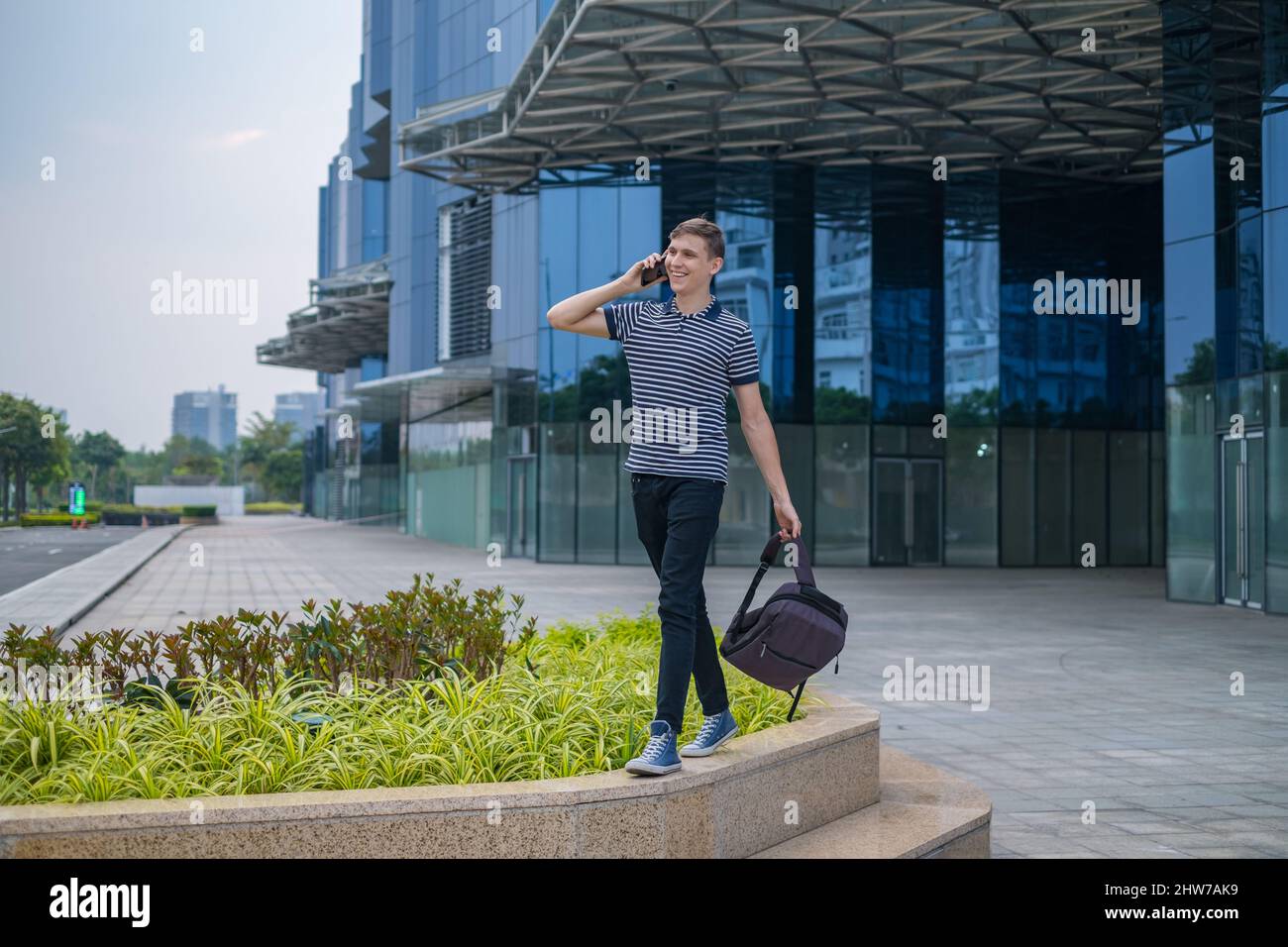 Giovane uomo caucasico in abbigliamento casual con lo zaino che cammina vicino a moderni edifici d'ufficio e fare telefonate. Persone che usano il telefono. Alta qualità Foto Stock