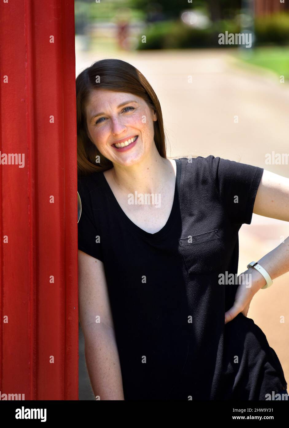 La giovane donna si appoggia contro una cabina telefonica rossa nel centro di Collierville, Tennessee. Ha una maglietta nera e sorride felicemente. Foto Stock