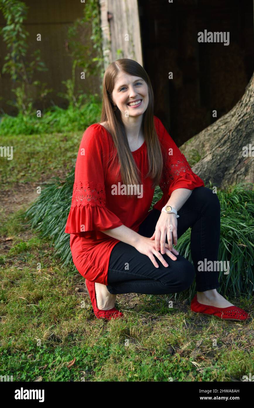 La giovane donna si inginocchia nel suo cortile e sorride. Si trova di fronte ad un albero e ad un granaio rustico di legno. Indossa una camicia rossa e dei jeans neri. Foto Stock