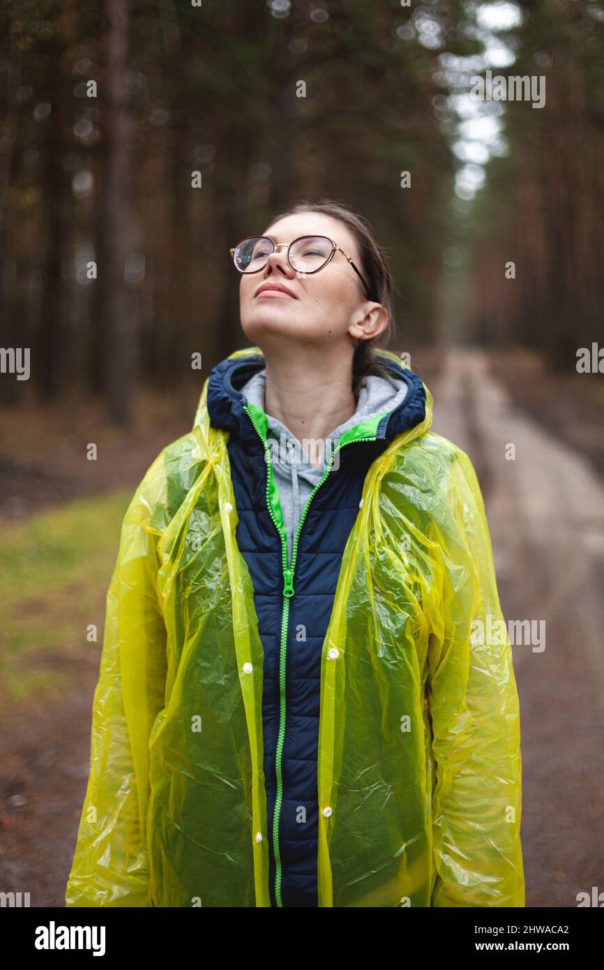 La giovane donna respira aria fresca nella foresta dopo la pioggia, riposa, rilassa, in piedi in un impermeabile giallo, respira ad occhi chiusi Foto Stock