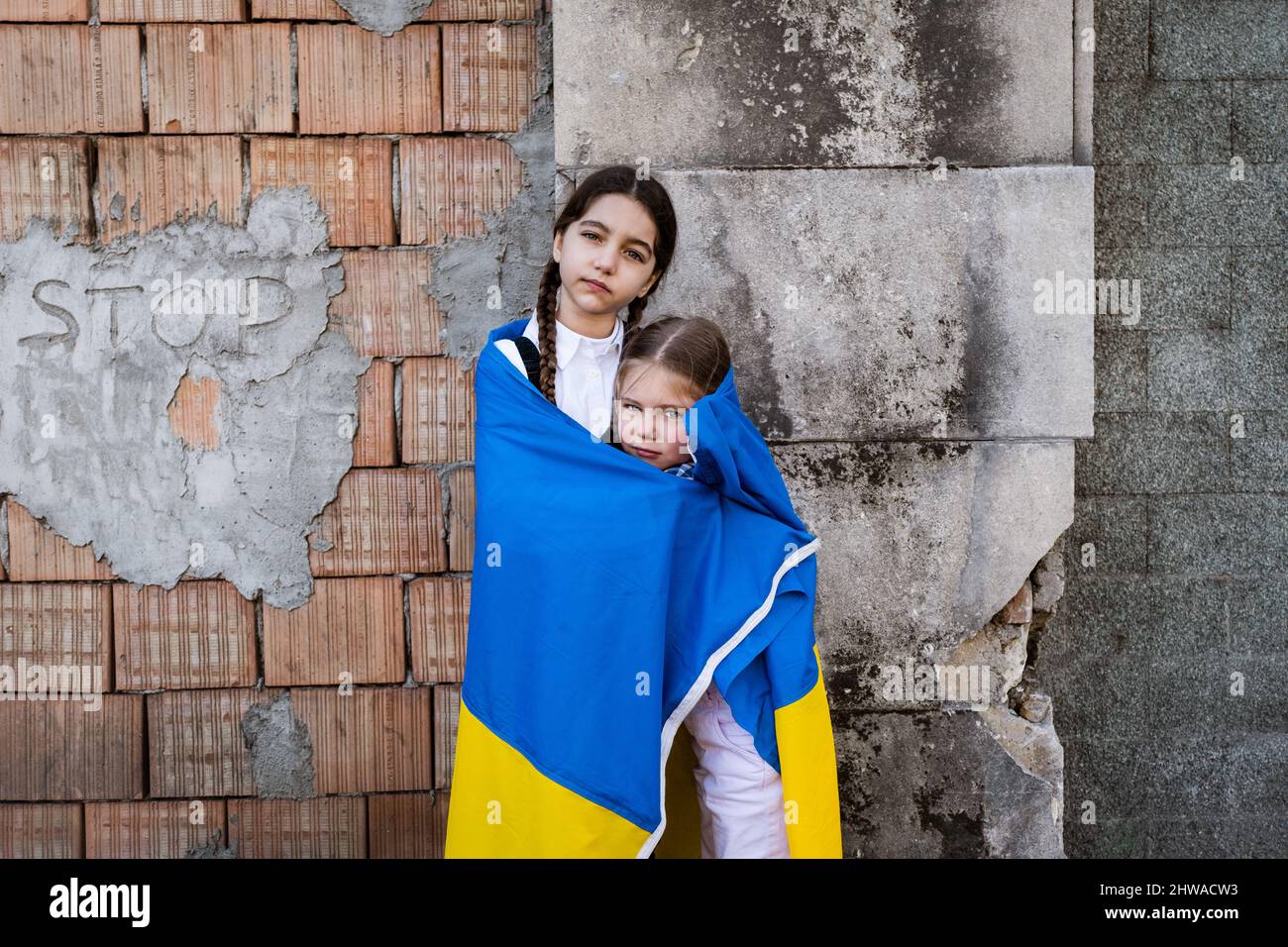 Bambine con bandiera Ucraina di fronte ad un muro distrutto dalle bombe. Le bambine ondano la bandiera nazionale mentre pregano per la pace Foto Stock