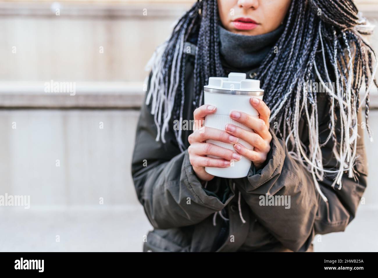 Taglia la donna irriconoscibile in un outerwear caldo con dreadlock che tiene thermos bianchi con caffè caldo nelle mani mentre si alza in strada Foto Stock