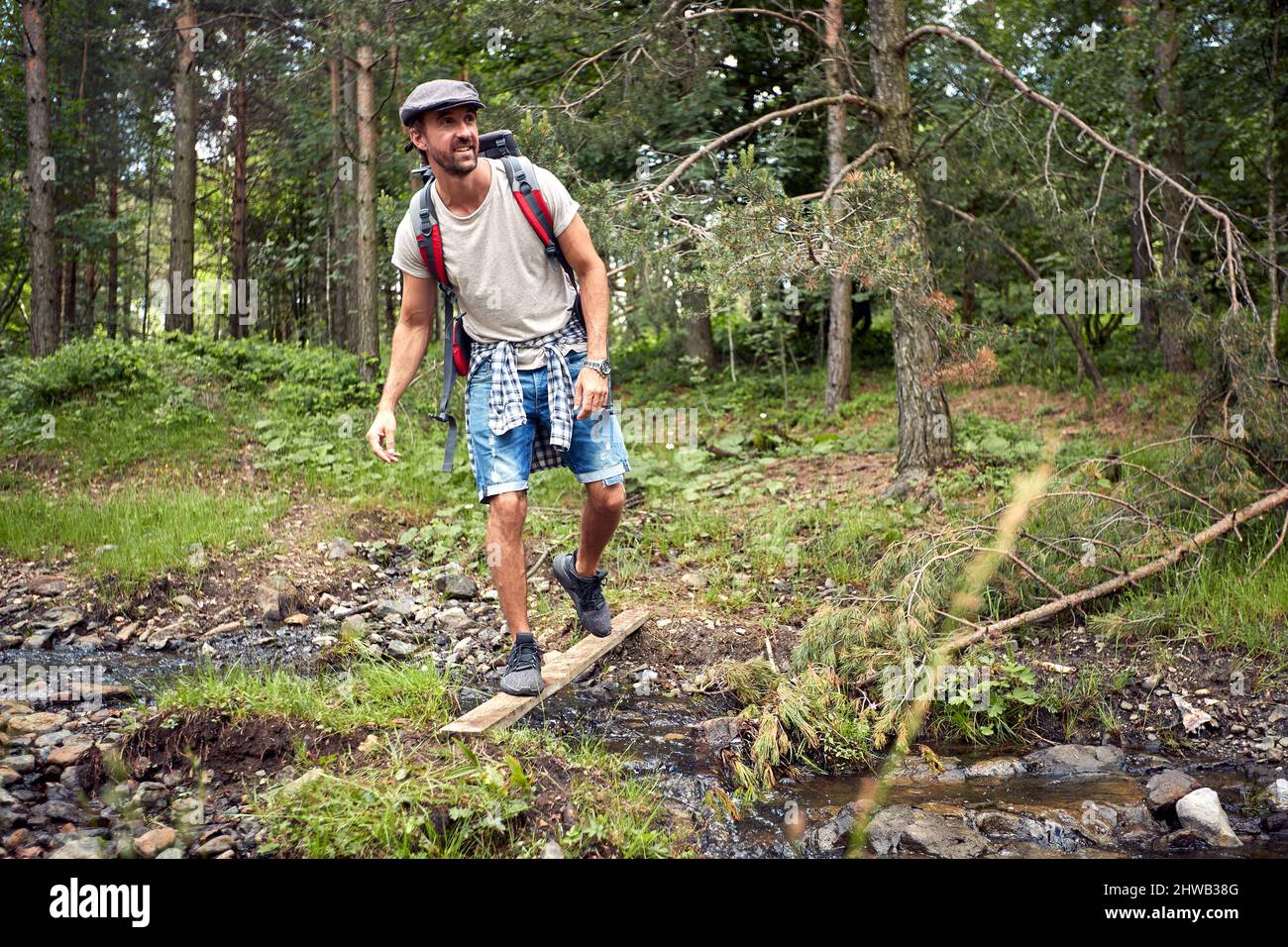 Giovane escursionista in cappello con zaino che attraversa un piccolo torrente nella foresta. Natura all'aperto. Sport, libertà, concetto di vacanza. Foto Stock