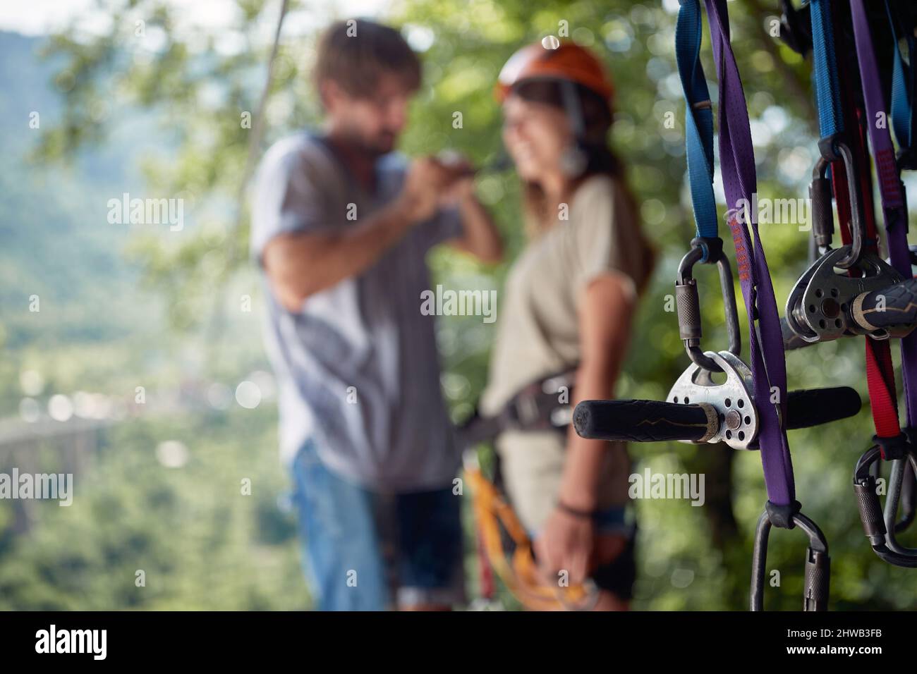 Fuoco selettivo sull'ingranaggio di protezione con sfondo sfocato della coppia. Giovane coppia attraente in abbigliamento casual che si prepara per l'avventura zip line in f Foto Stock