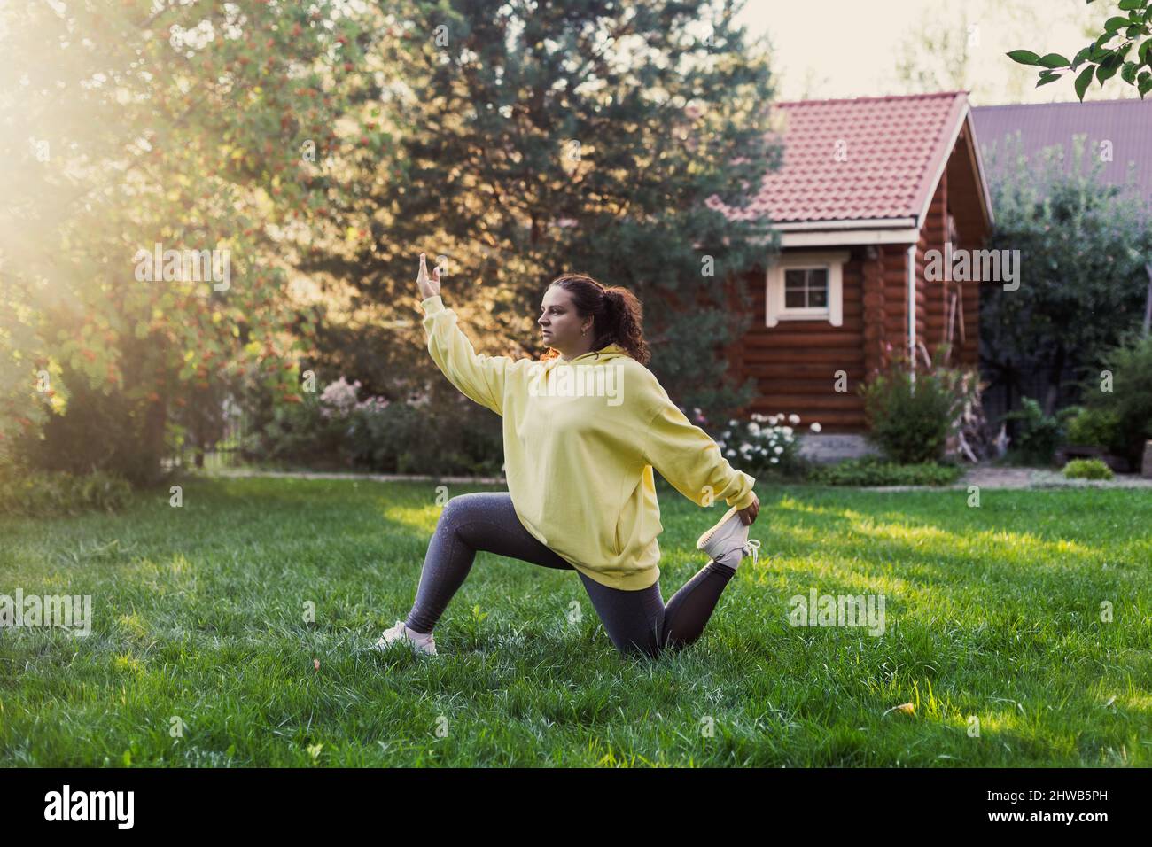 Giovane donna che indossa abiti sportivi facendo esercizi di yoga sul cortile sul prato verde con casa di campagna in legno e alti alberi sullo sfondo. Corpo Foto Stock