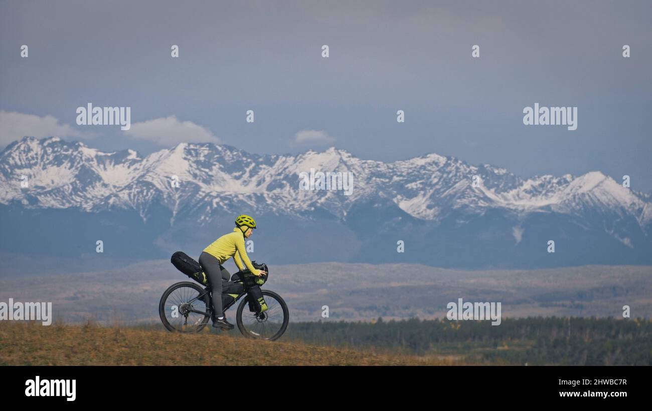 La donna viaggia su percorsi ciclabili misti con bikepacking. Il viaggio  del viaggiatore con le borse della bicicletta. Sport turismo bikepacking,  bici, abbigliamento sportivo in verde nero. Montagna innevata Foto stock -