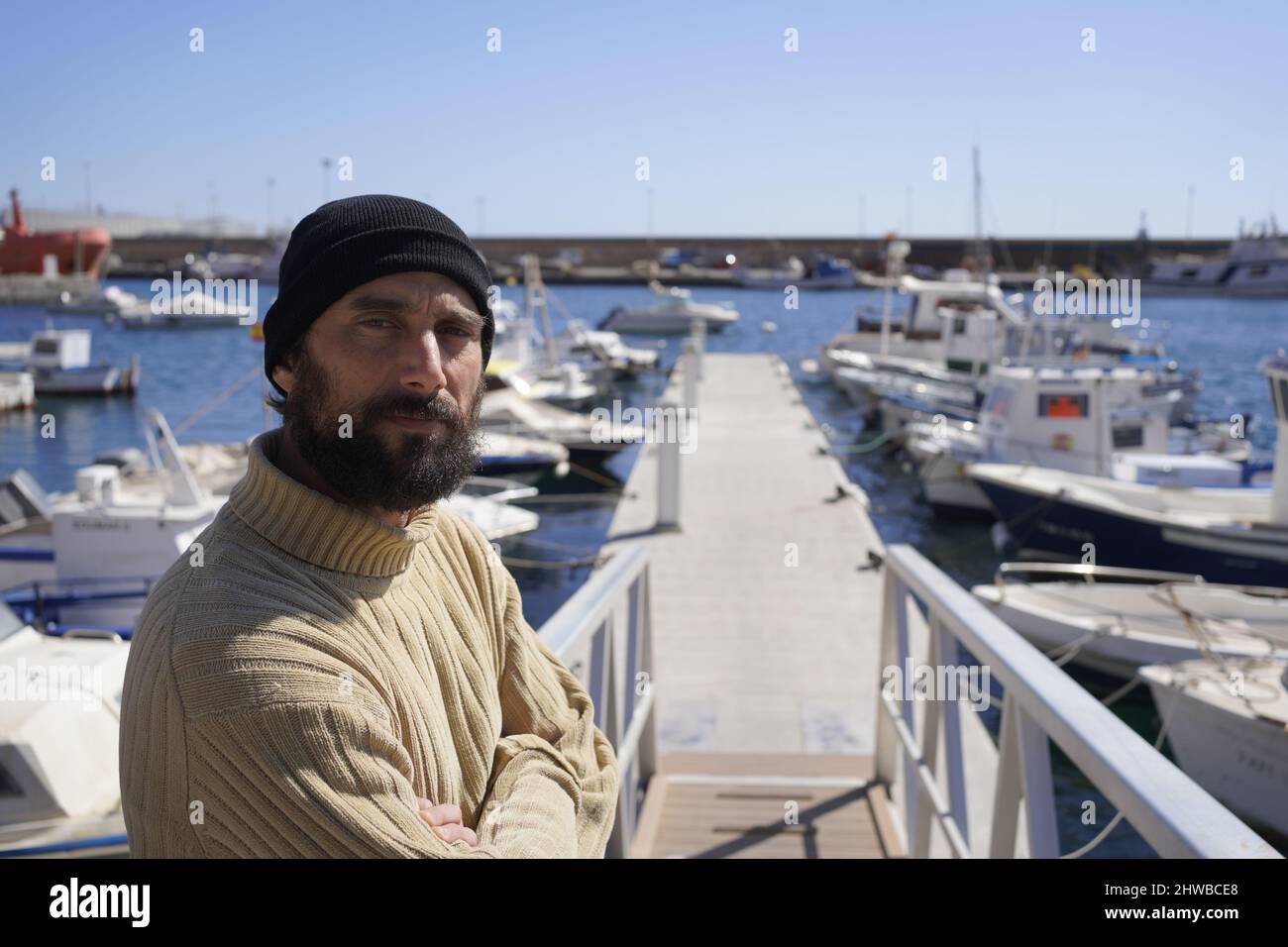 Un giovane marinaio alto e robusto con barba in berretto di lana e pullover a collo alto è in piedi su una nave in un porto marittimo Foto Stock