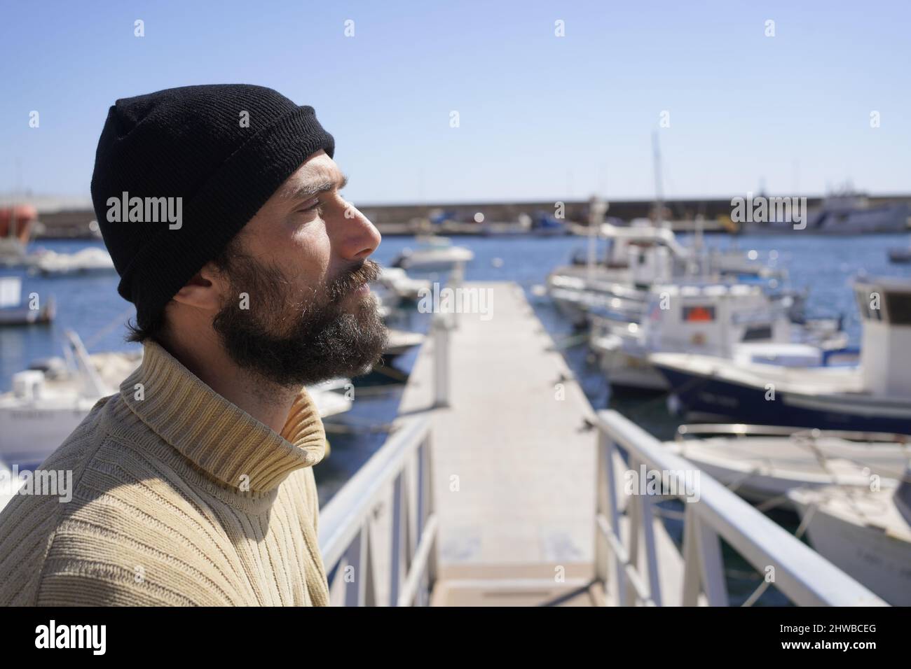 Un giovane marinaio alto e robusto con barba in berretto di lana e pullover a collo alto è in piedi su una nave in un porto marittimo Foto Stock