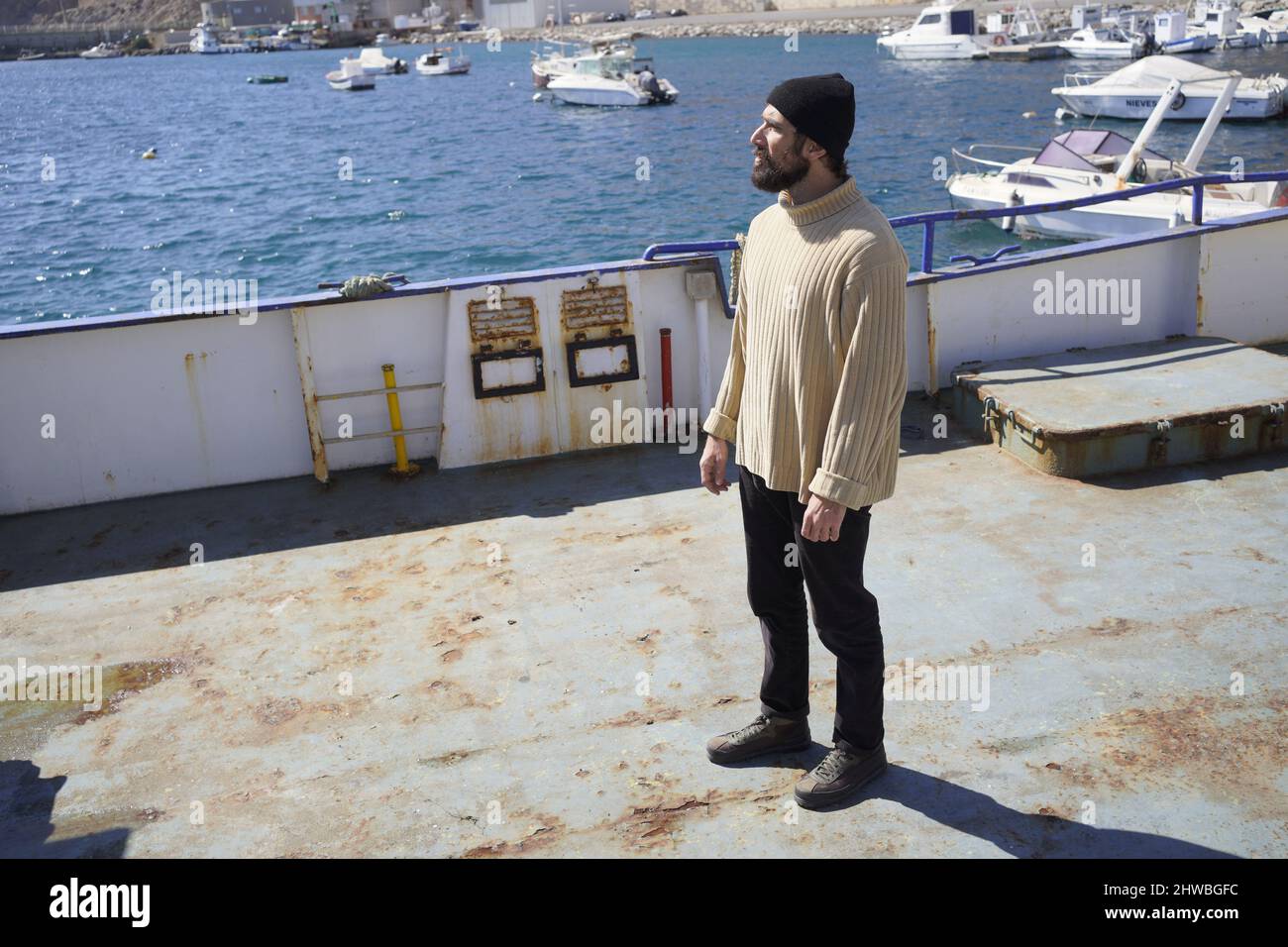 Un giovane marinaio alto e robusto con barba in berretto di lana e pullover a collo alto è in piedi su una nave in un porto marittimo Foto Stock