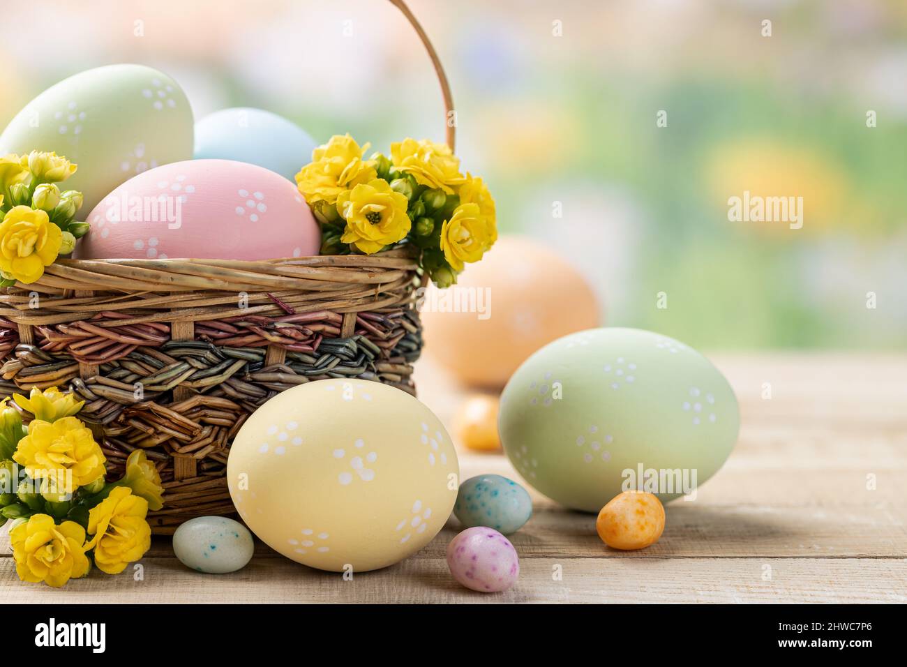 Uova di pasqua colorate con cestino, caramelle e fiori su un tavolo di legno con sfondo primavera colorato Foto Stock