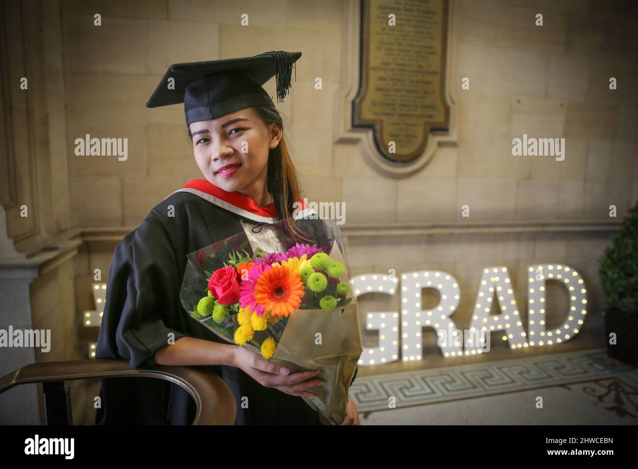 Una graziosa laureata asiatica si pone con fiori per le fotografie alla sua cerimonia di laurea. Questo evento formale in cui i diplomi (prima) cambiano Foto Stock